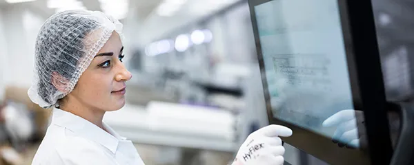 Berufserfahrene-lts Eine Frau mit weißem Kittel steht in einer Produktionshalle an einem Monitor.