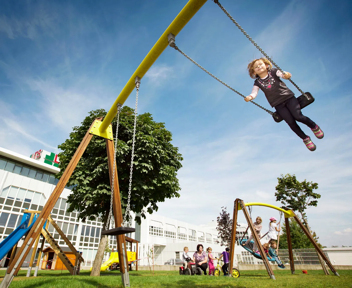 Mehrere Kinder schaukeln auf dem Rasen-Spielplatz des LTS-Kindergartens, weitere Kinder stehen bei einer Erzieherin; über dem Bild liegt der Text ‚Betriebseigener Kindergarten‘.