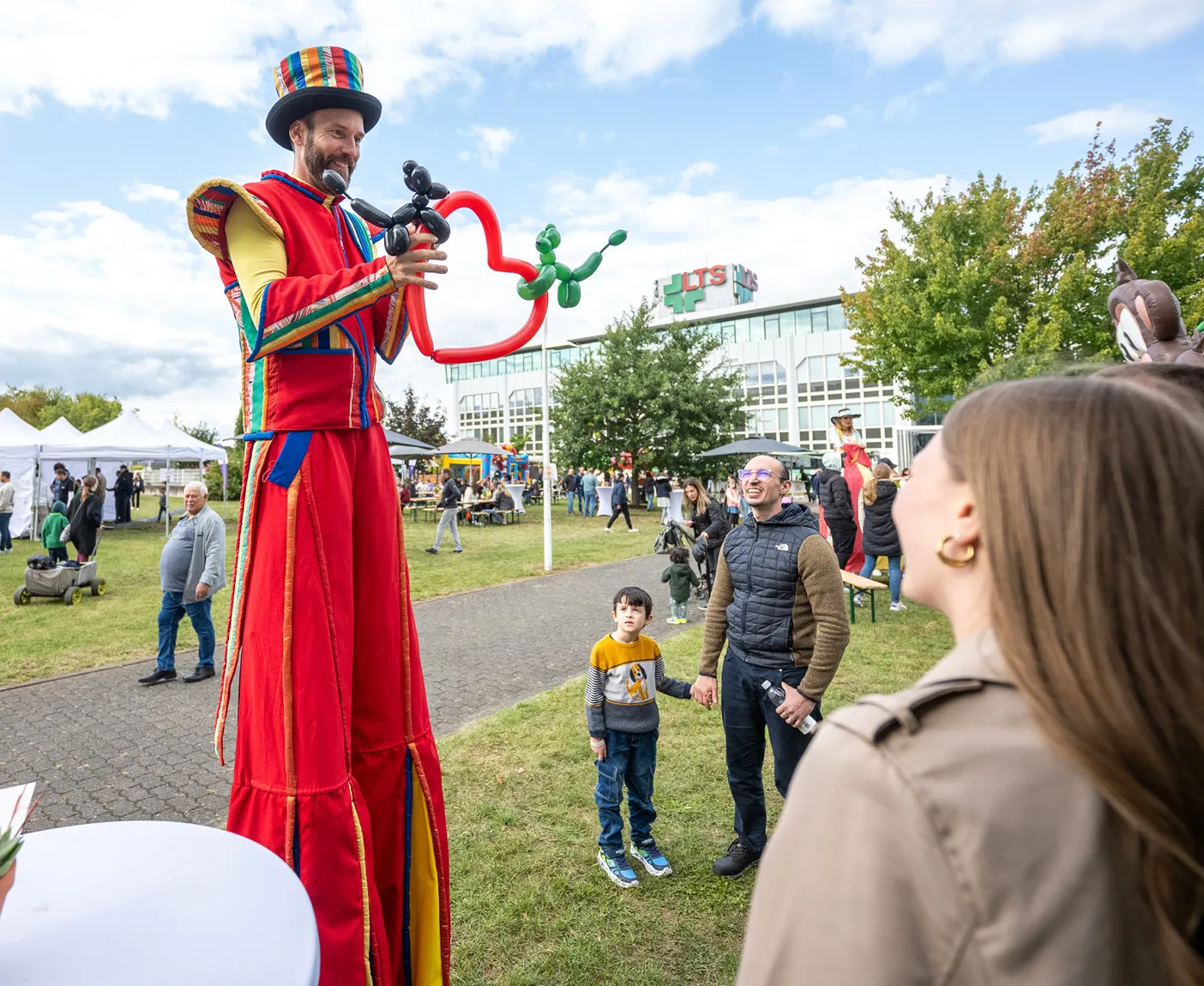 Ein rot gekleideter Luftballonkünstler auf Stelzen formt vor Zuschauern ein Herz aus Ballons; über dem Bild liegt der Text ‚Familienfest‘.