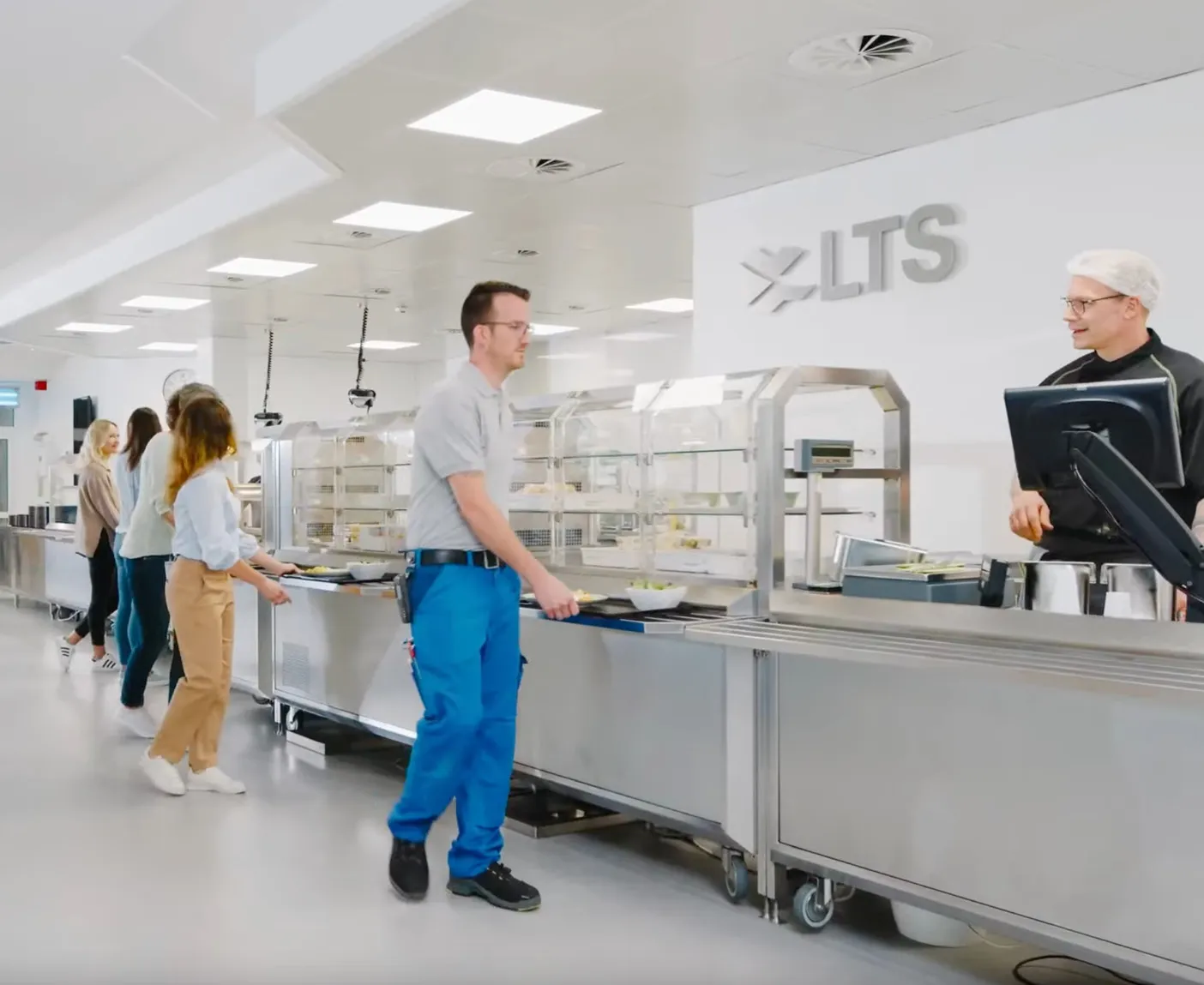 LTS-Mitarbeitende stehen in der LTS-Kantine an der Essensausgabe mit Tabletts in der Hand; über dem Bild liegt der Text ‚LTS Kantine‘.