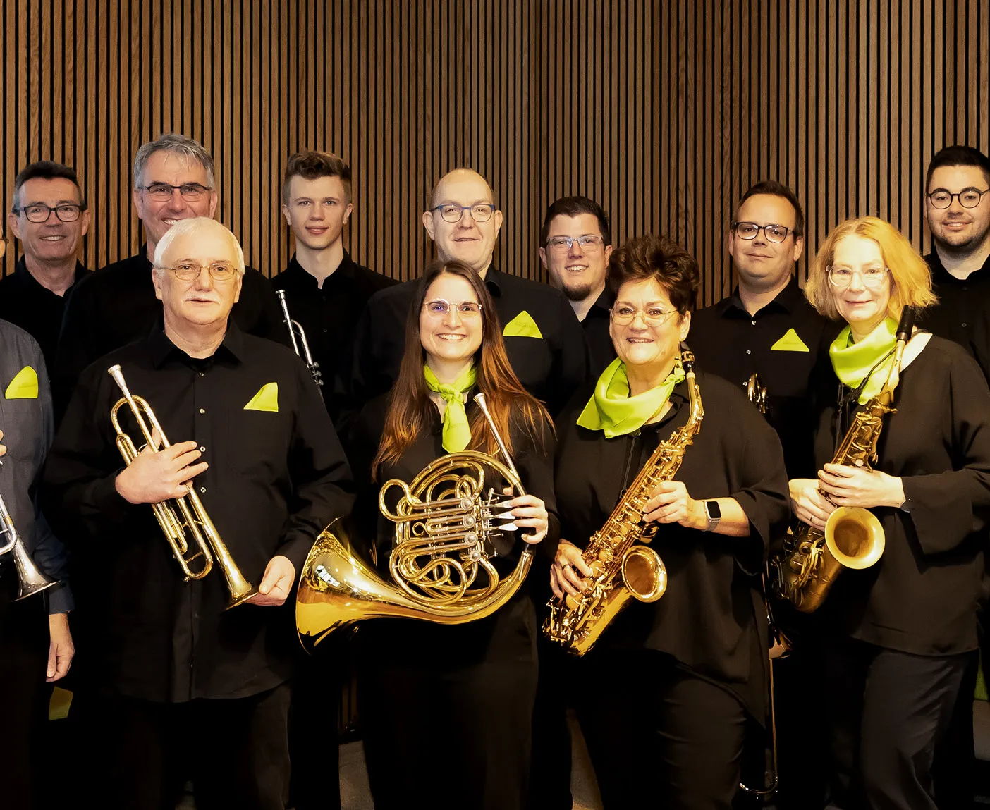 Mitglieder des LTS-Orchesters in schwarzer Kleidung mit gelben Halstüchern mit Blasinstrumenten; über dem Bild liegt der Text ‚LTS-Orchester‘.