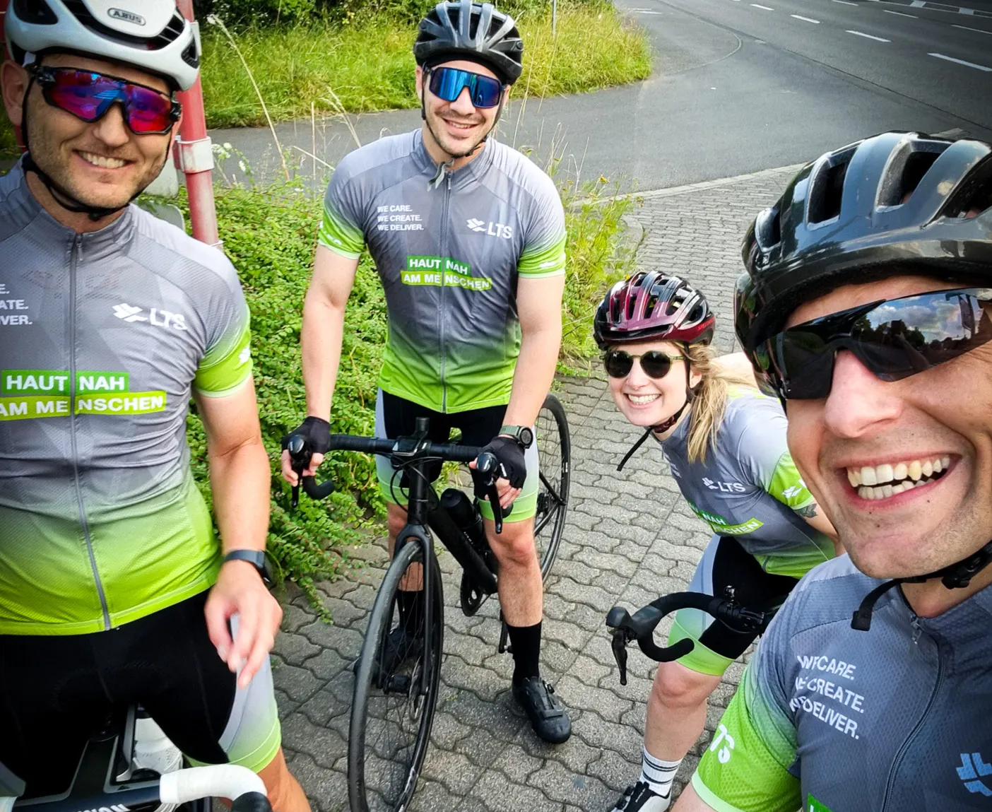 Vier Radfahrer:innen in grau-grünen LTS-Trikots stehen mit ihren Rennrädern am Straßenrand; über dem Bild liegt der Text ‚LTS-Rennradgruppe‘.