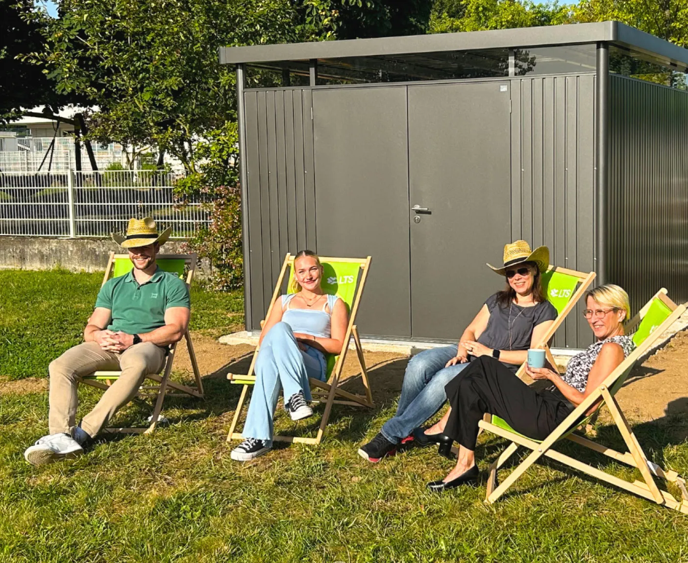 Vier LTS-Mitarbeiter:innen entspannen in Liegestühlen auf einer Wiese mit Gartenhaus; über dem Bild liegt der Text ‚Mittagspause im Grünen‘.