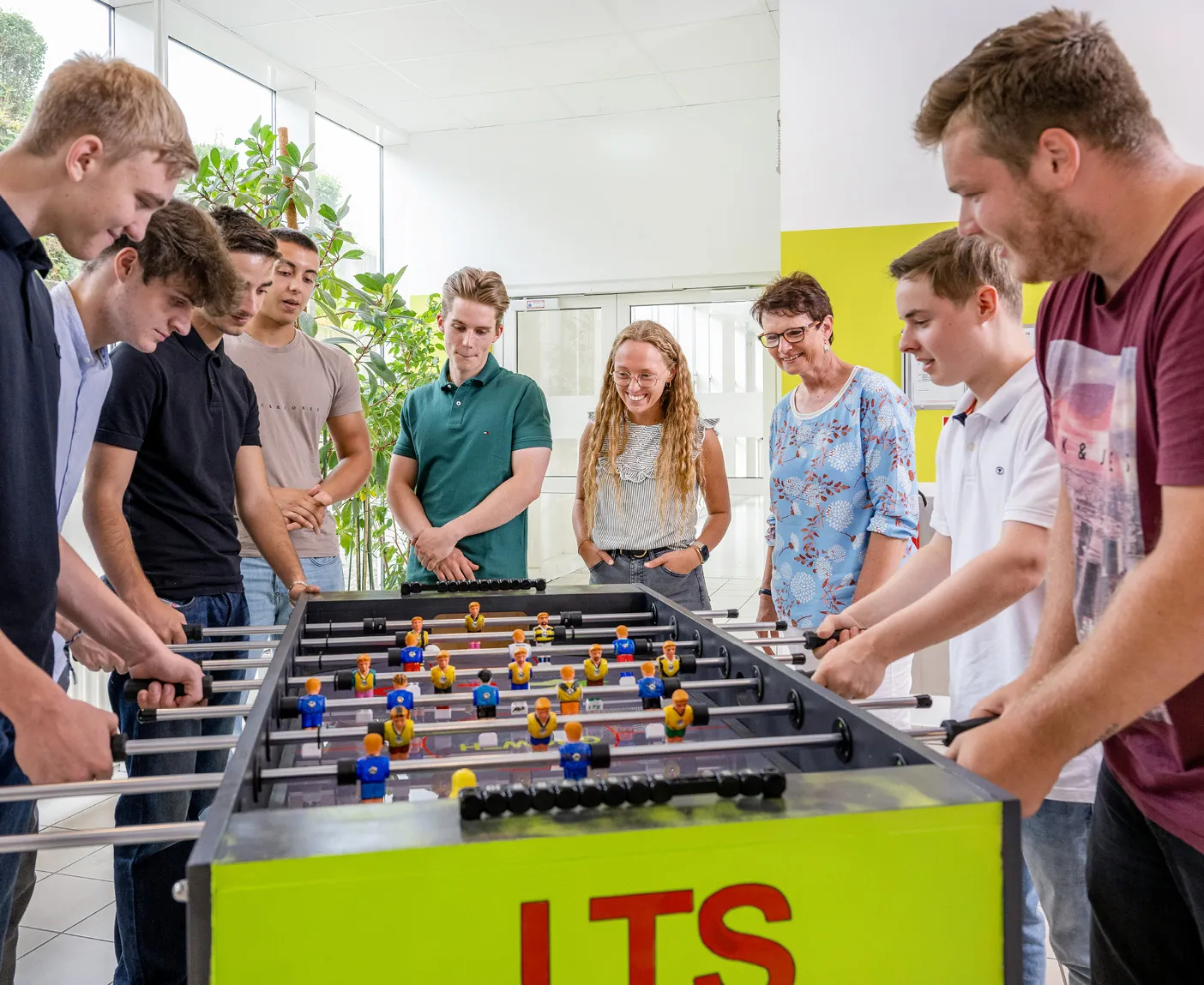 Mehrere LTS-Mitarbeiter:innen spielen in einem hellen Raum gemeinsam an einem Kicker-Tisch mit der Aufschrift ‚LTS‘; über dem Bild liegt der Text ‚Pausenspaß und -spiel‘.