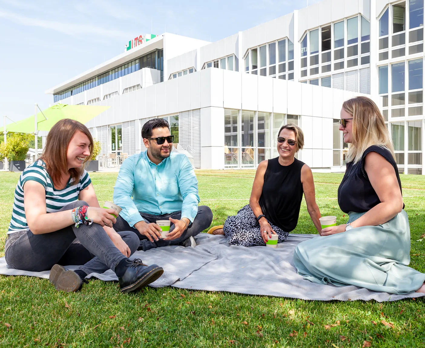 Vier LTS-Mitarbeitende sitzen entspannt auf einer Picknickdecke vor dem LTS-Gebäude; über dem Bild liegt der Text ‚Relaxen im Grünen‘.