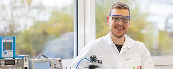 Ein junger Mann im Laborkittel steht in einem LTS-Labor neben technischen Geräten.