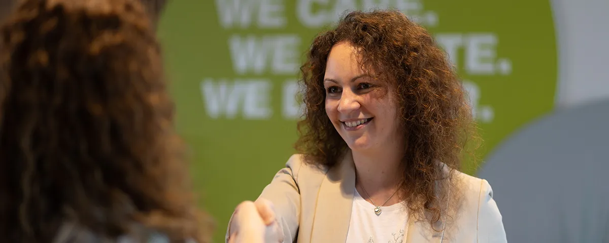 Eine LTS-Mitarbeiterin mit lockigem Haar und weißer Bluse reicht die Hand zum Handschlag. Im Hintergrund steht weiß auf grünem Hintergrund der LTS-Slogan: we care, we create, we deliver.